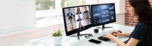 Young woman on online pbx voip video conference with nine other business men and women. Second monitor displays infographics and metrics.