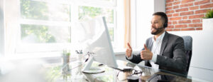 Customer service agent bearded man on video call with client, giving two thumbs up to camera. Business man is smiling and wearing a headset. VOIP connection.