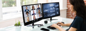 Young woman on online pbx voip video conference with nine other business men and women. Second monitor displays infographics and metrics.