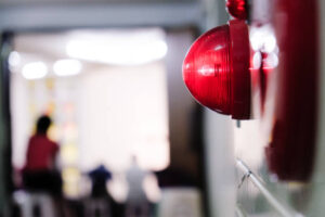 Red alarm on side of brick wall that is lit up. Concept of fire, water, warning, danger, building, fire alarm. People silhouetted in background leaving building.