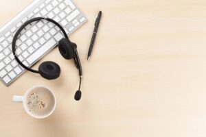Top-down view of Mac keyboard on bright wooden desk with telecommunications pbx voip headset resting on top of it. Black ballpoint pen and mug of coffee rest on desk.