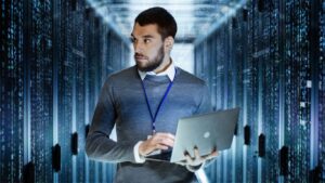 Man with laptop and badge in middle of data center hallway, monitoring data cabinets. Server cabinets are wrapped in code.