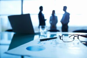 Foreground shows pair of glasses and pen on metric graph paper. In background lies three silhouetted business people.