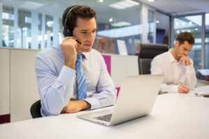 Two men at conference room table: one on video call with headset on. Second business man pensively writing down notes in modern office environment.