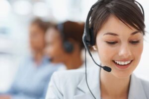 Young woman in call center or service desk smiling and doing work. In background are the silhouettes of two other women are also on calls