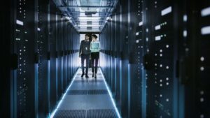 Man and woman walking down hallway in working data center. Floor and server cabinets are illuminated by light. Monitoring data center.