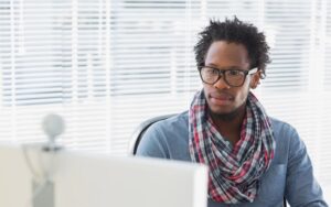 Young trendy man with glasses and scarf on video conference call. Background is window with natural sunlight coming through. Webcam is visible on top of computer monitor.