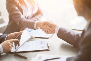 Image of handshake with third person on mobile tablet or device. Studying finances or other statistics. Natural sunlight illuminates room.