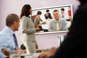 Image of three people on video conference call over the internet having a meeting. Video is displayed on TV. Four business people on opposite end of call smiling.